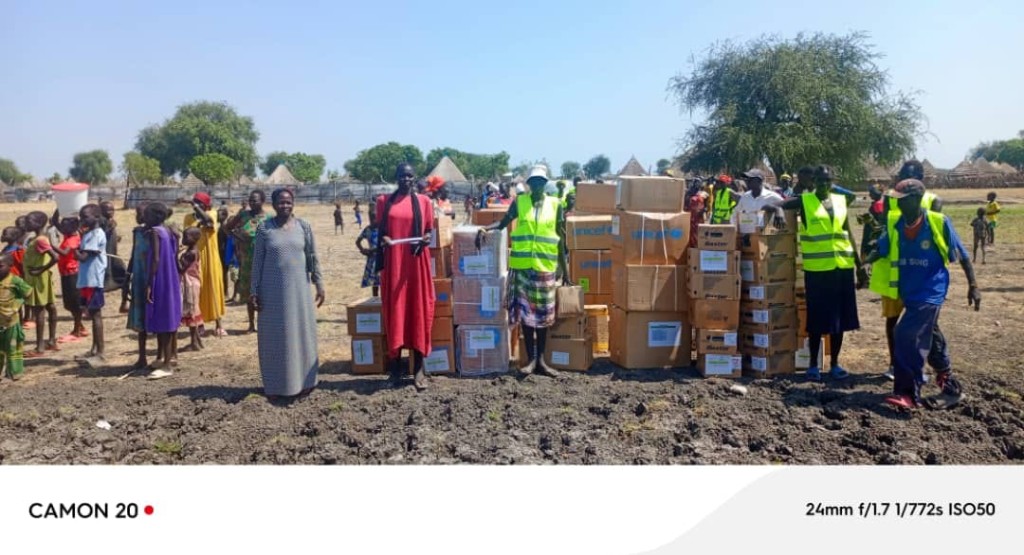 Women, children and aid workers gathered around stacked humanitarian supply boxes in a rural community.